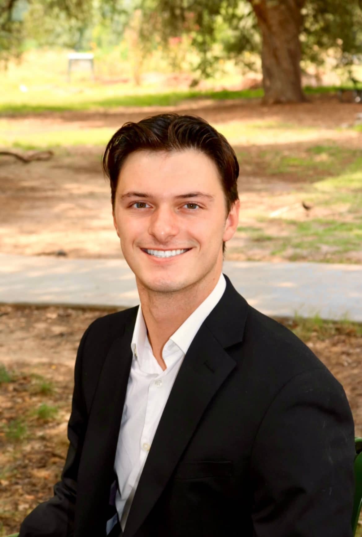 Luke Allen, photographed outdoors under live oaks in a dark jacket and open-collar white shirt.